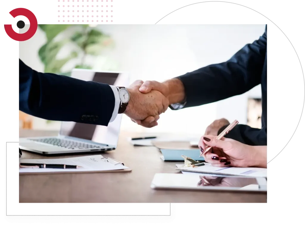 In this close-up photo, two business men are seated at a table across from each other. They are both wearing formal suits, which convey a sense of professionalism. In the center of the image, their hands are clasped together in a firm handshake, symbolizing a business agreement or partnership. The table is made of polished wood and is strewn with documents and pens, suggesting a serious business meeting. The background is softly blurred, keeping the focus on the handshake. Overall, this image captures the essence of a formal business interaction and partnership.